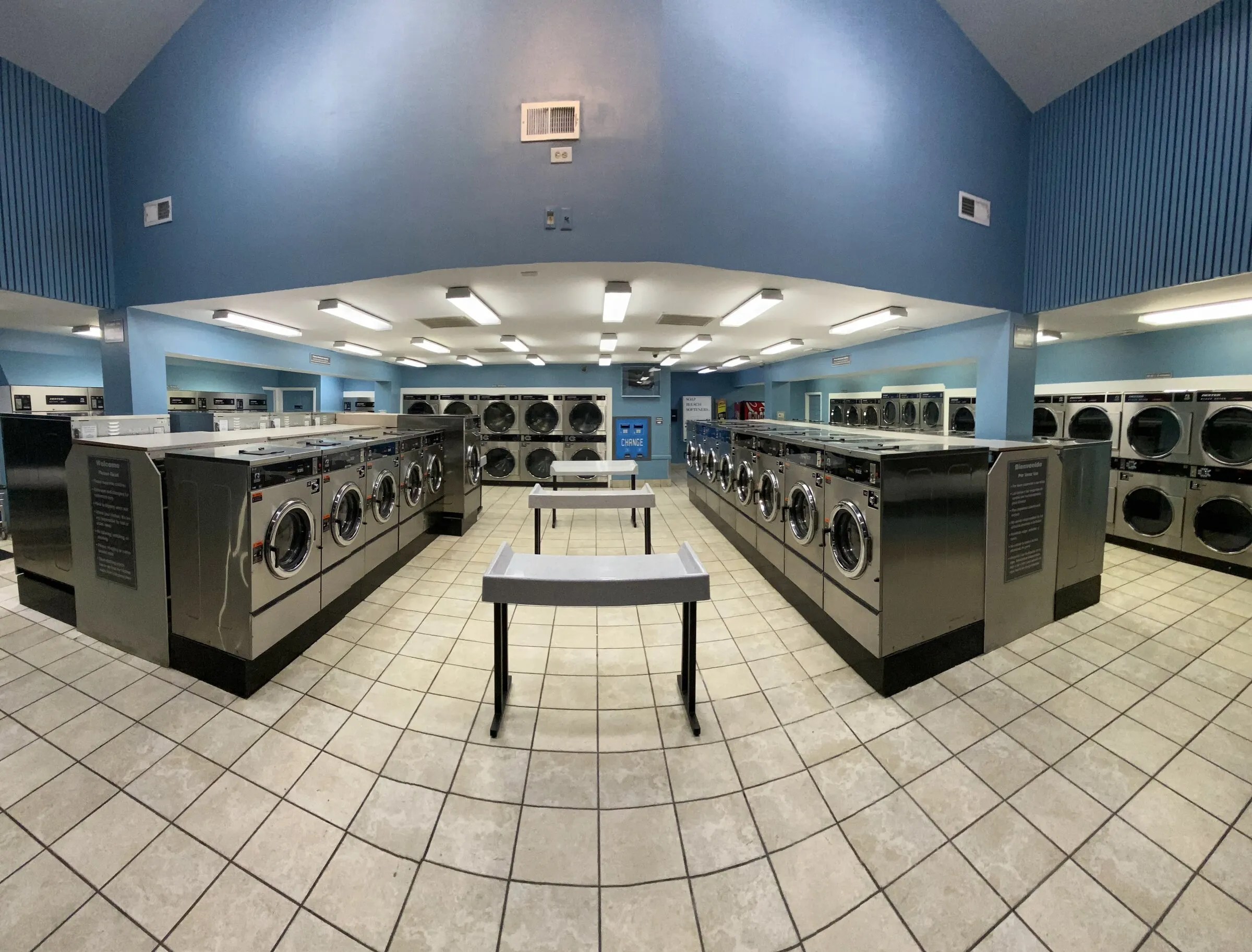 The main floor of the laundromat: two rows of front-load washers facing each other with stainless folding tables between them and dryers along the back wall.