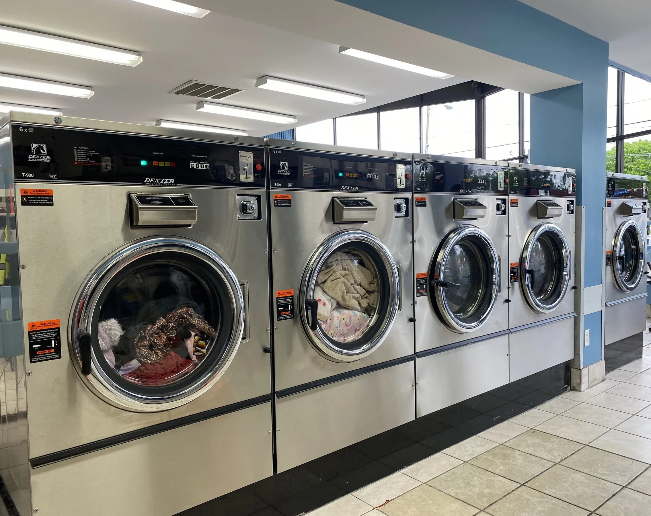 60-pound washers in a row, some running with clothes and a comforter inside, daylight from the windows behind.