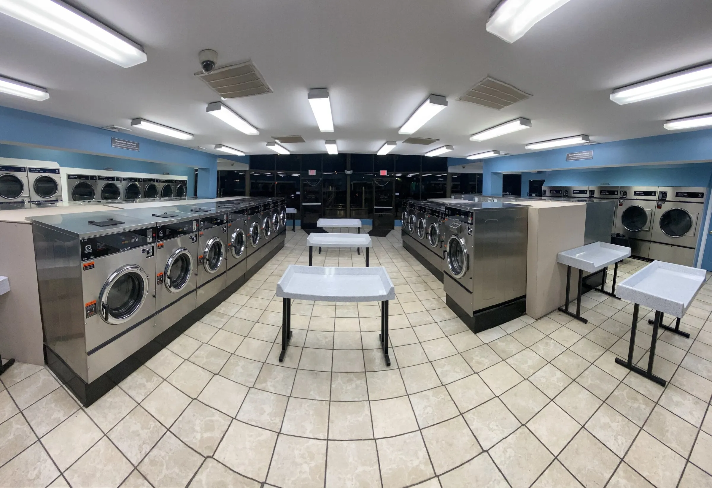 Three stainless folding tables between washer islands, dryers on both sides and a security camera on the ceiling.